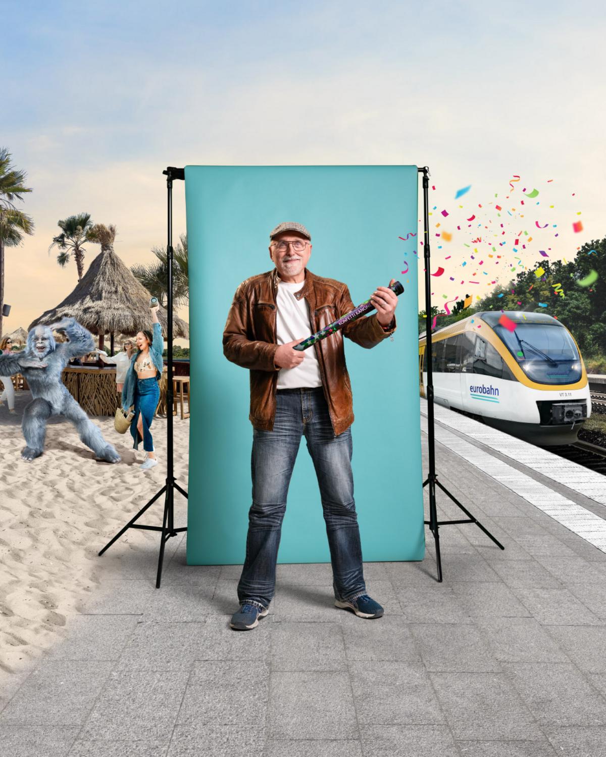 Alfons am Bahnsteig in Münster Ein älterer Herr in brauner Lederjacke und mit Schiebermütze hält eine Konfettikanone in der Hand, die er gerade abgefeuert hat und steht am Bahnsteig in Münster vor einer türkisen Leinwand. Links ist ein Beachclub mit feiernden Leuten zu sehen sowie ein tanzender Mensch im Yeti-Kostüm und rechts ein gelb-weißer Zug der Linie RB 69 von der eurobahn.