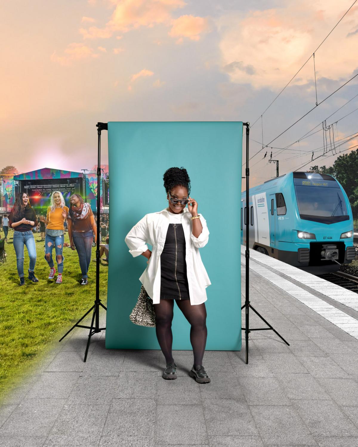 Arieth am Bahnsteig in Hengelo Eine Frau in einem schwarz-weißen Outfit steht am Bahnsteig in Hengelo vor einer türkisen Leinwand. Links ist eine Konzertbühne mit eine Menge von Konzertbesuchern zu sehen und rechts ein gelb-weißer Zug der Linie RB 61 von der eurobahn.