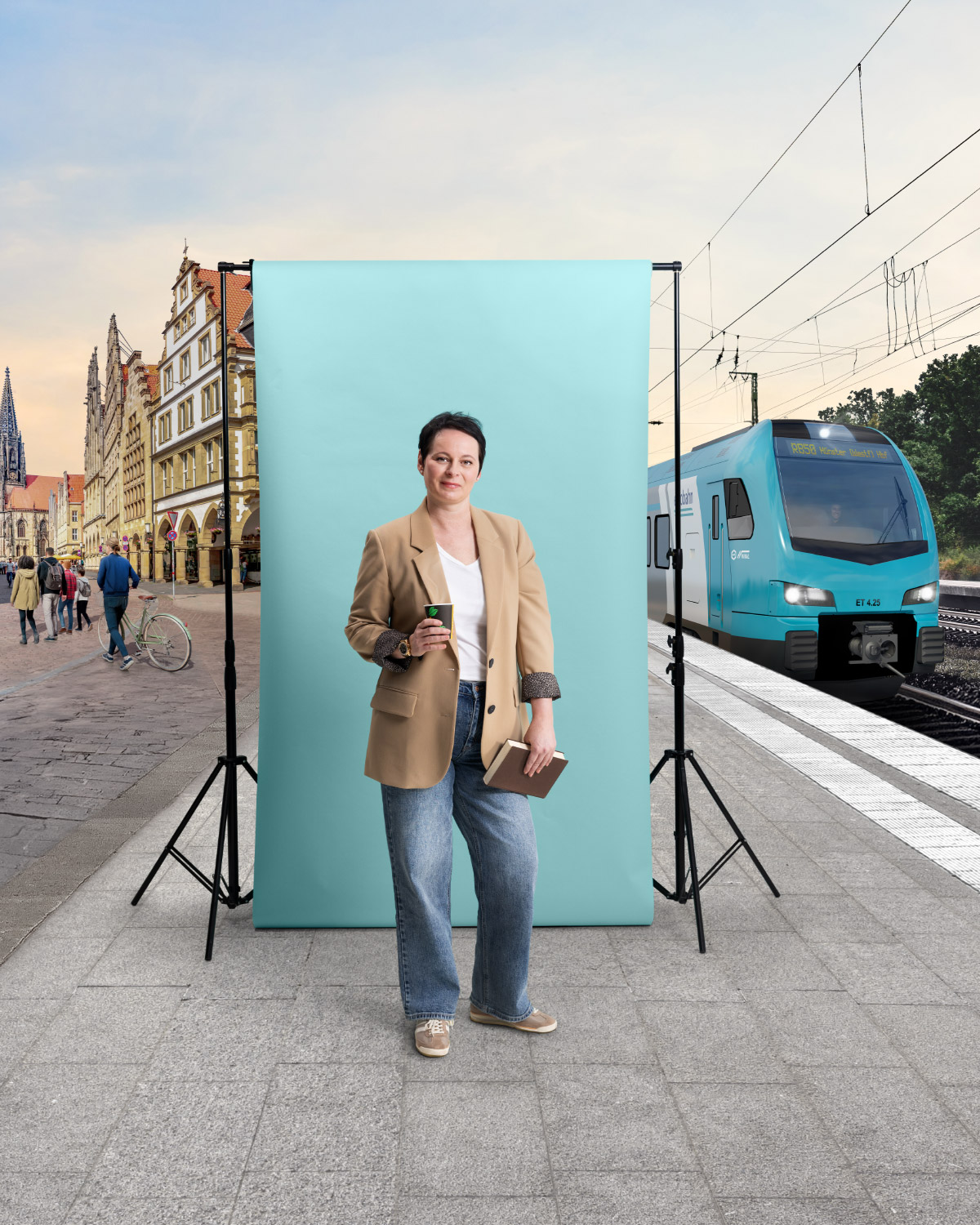 Eine mittelalte Frau in beigem Blazer und Jeans hält in der einen Hand ein Buch, in der anderen einen Kaffeebecher. Sie steht am Bahnsteig in Münster vor einer türkisen Leinwand. Links ist die Altstadt zu sehen und rechts ein türkiser Zug der Linie RB 50 von der eurobahn.