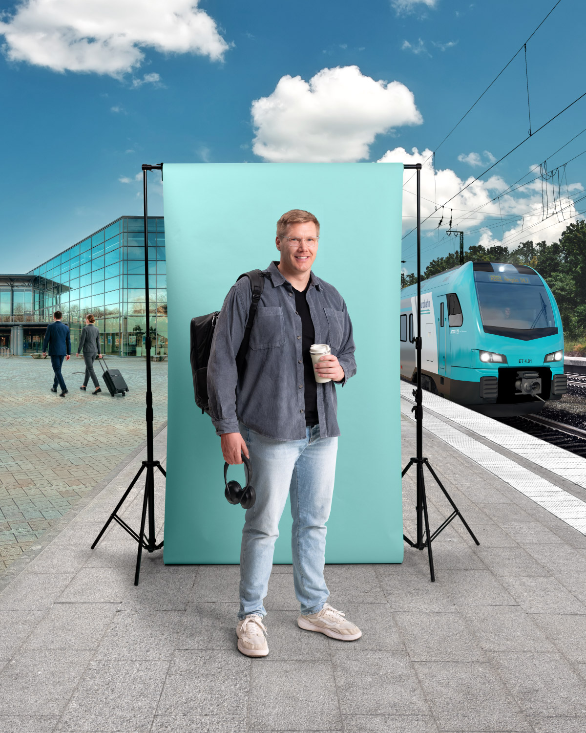 Ein mittelalter Mann in Jeansjacke und mit Rucksack hält in der einen Hand einen Kopfhörer, in der anderen einen Kaffeebecher. Er steht am Bahnsteig in Osnabrück vor einer türkisen Leinwand. Links ist ein modernes Gebäude mit Glasfront zu sehen und rechts ein türkiser Zug der Linie RB 61 von der eurobahn.