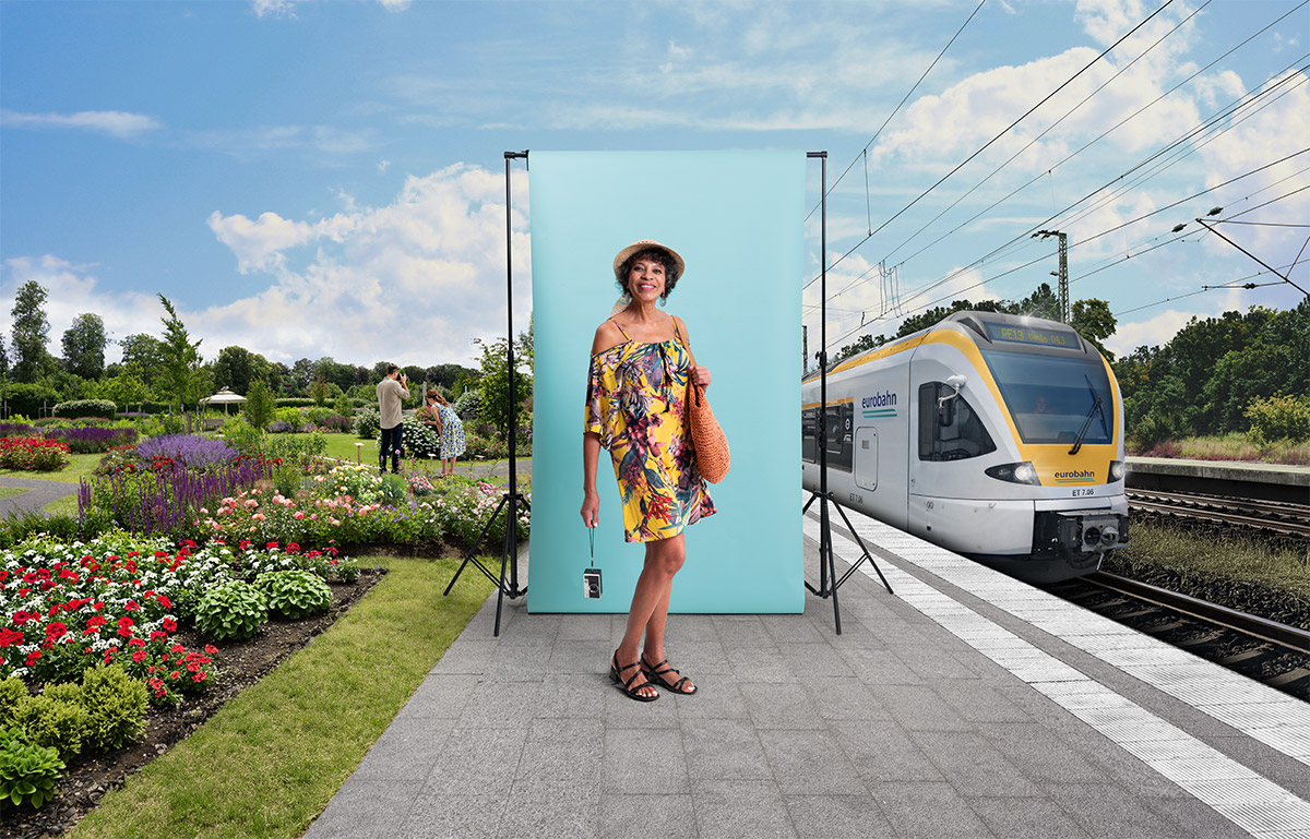 Eine ältere Dame einem bunten Sommerkleid steht am Bahnsteig in Neuss vor einer türkisen Leinwand. Links ist die Landesgartenschau Neuss zu sehen und rechts ein gelb-weißer Zug der Linie RE 13 von der eurobahn.