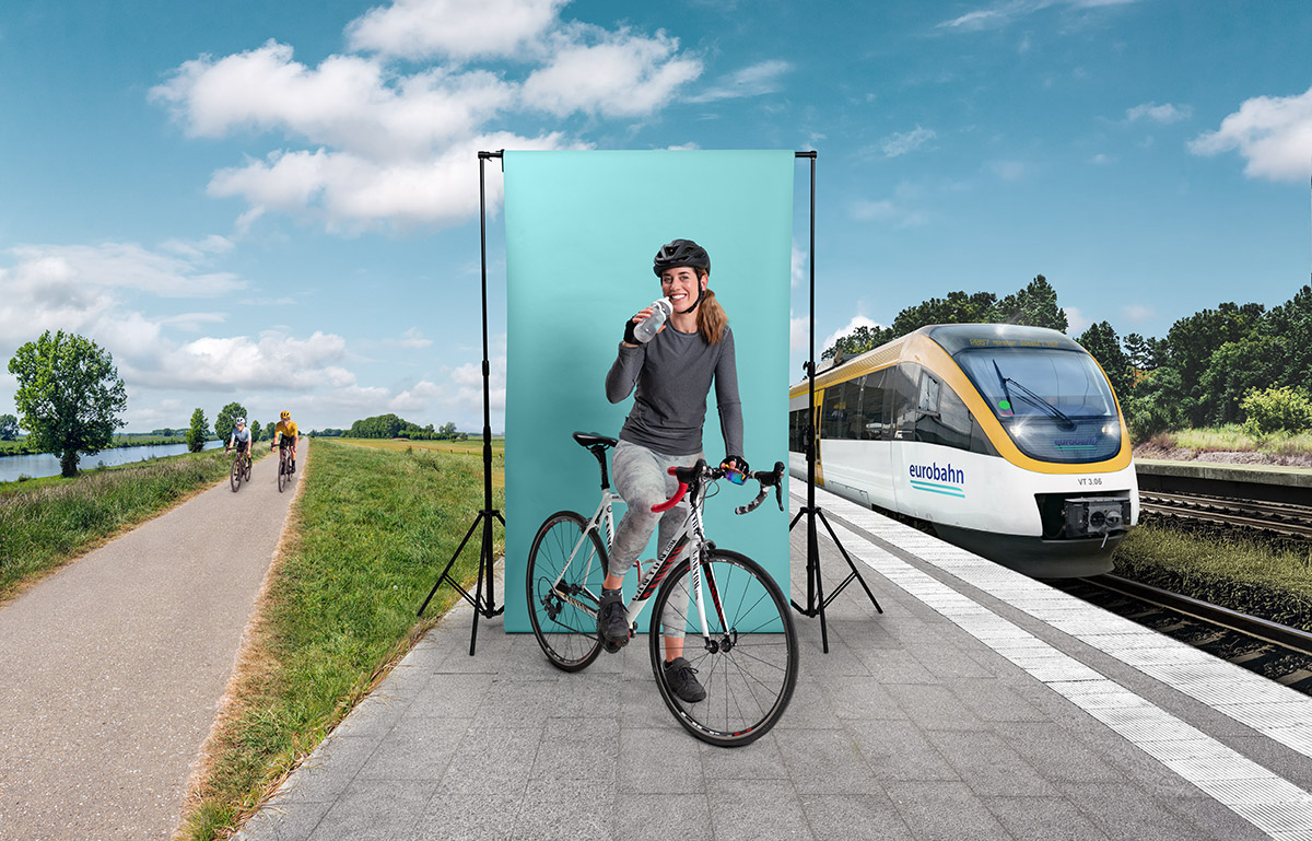 Eine Frau in Sportkleidung und Fahrradhelm sitzt auf einem Rennrad vor einer türkisen Leinwand und trinkt aus einer Flasche. Links sind zwei Fahrradfahrer auf einem Radweg in grüner Landschaft zu sehen und rechts ein Zug der eurobahn. Die Szene spielt an einem Bahnsteig bei sonnigem Wetter.