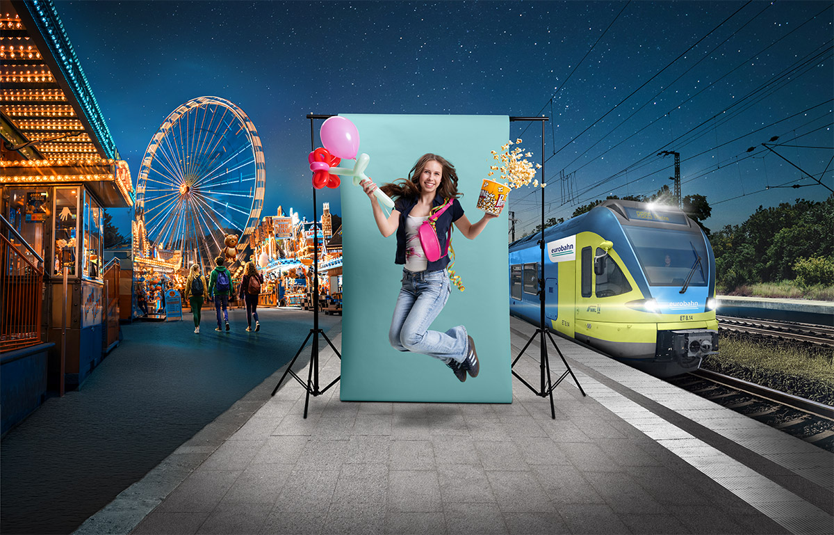 Eine junge Frau in Jeans und dunkelblauer Bluse springt vor einer türkisen Leinwand in die Luft. In der einen Hand hält sie eine Ballonfigur, in der anderen einen Becher Popcorn. Links ist eine beleuchtete Kirmes mit Riesenrad zu sehen und rechts ein Zug der eurobahn. Die Szene spielt an einem Bahnsteig bei Nacht unter einem sternenklaren Himmel.