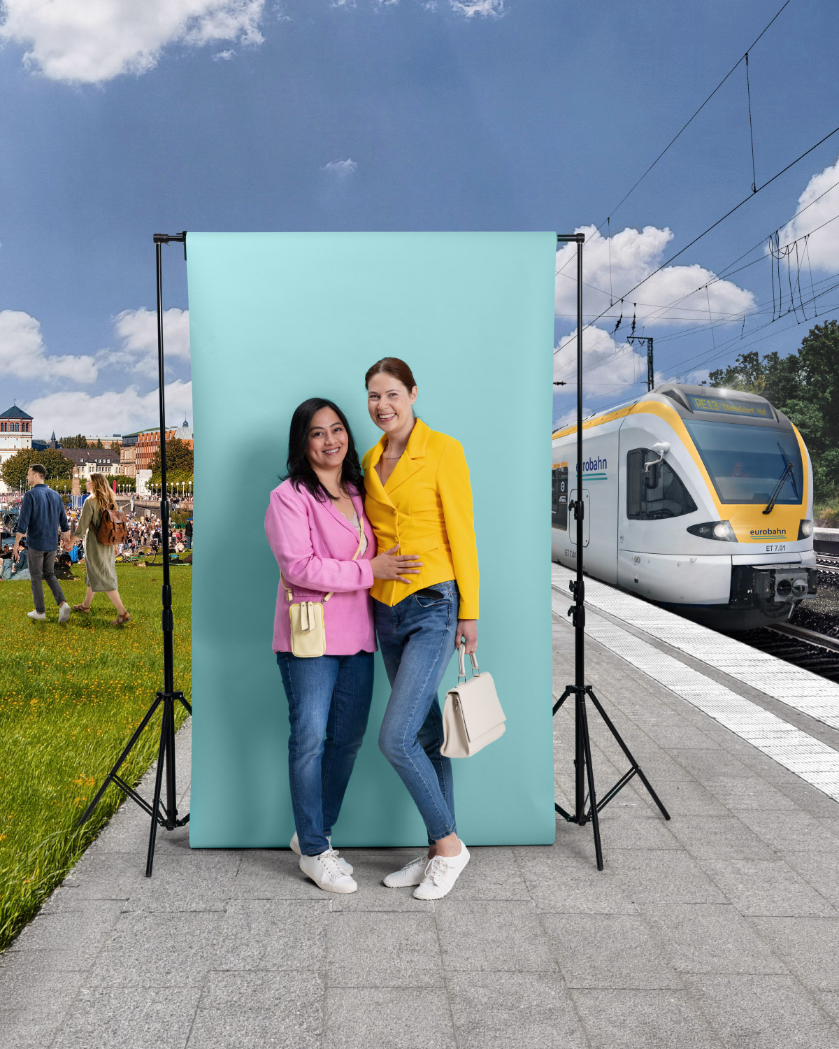 Zwei Frauen in farbenfrohen Blazern und Jeans stehen als Paar eng beieinander vor einer türkisen Leinwand und lächeln in die Kamera. Links ist eine belebte Grünfläche am Rhein in Düsseldorf zu sehen und rechts ein Zug der eurobahn. Die Szene spielt an einem Bahnsteig bei sonnigem Wetter.