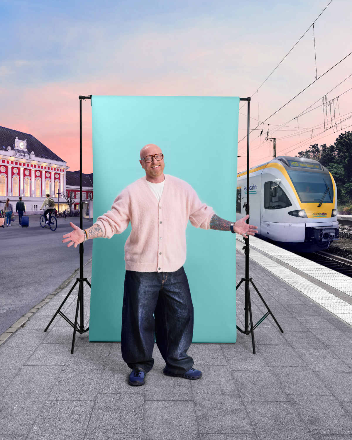 Ein Mann mit Brille, rosa Strickjacke und weiter Jeans steht mit ausgebreiteten Armen vor einer türkisen Leinwand und lächelt in die Kamera. Links ist der belebte Bahnhofsvorplatz von Hamm zu sehen und rechts ein Zug der eurobahn. Die Szene spielt an einem Bahnsteig vor einem rosa-blau gefärbten Abendhimmel.