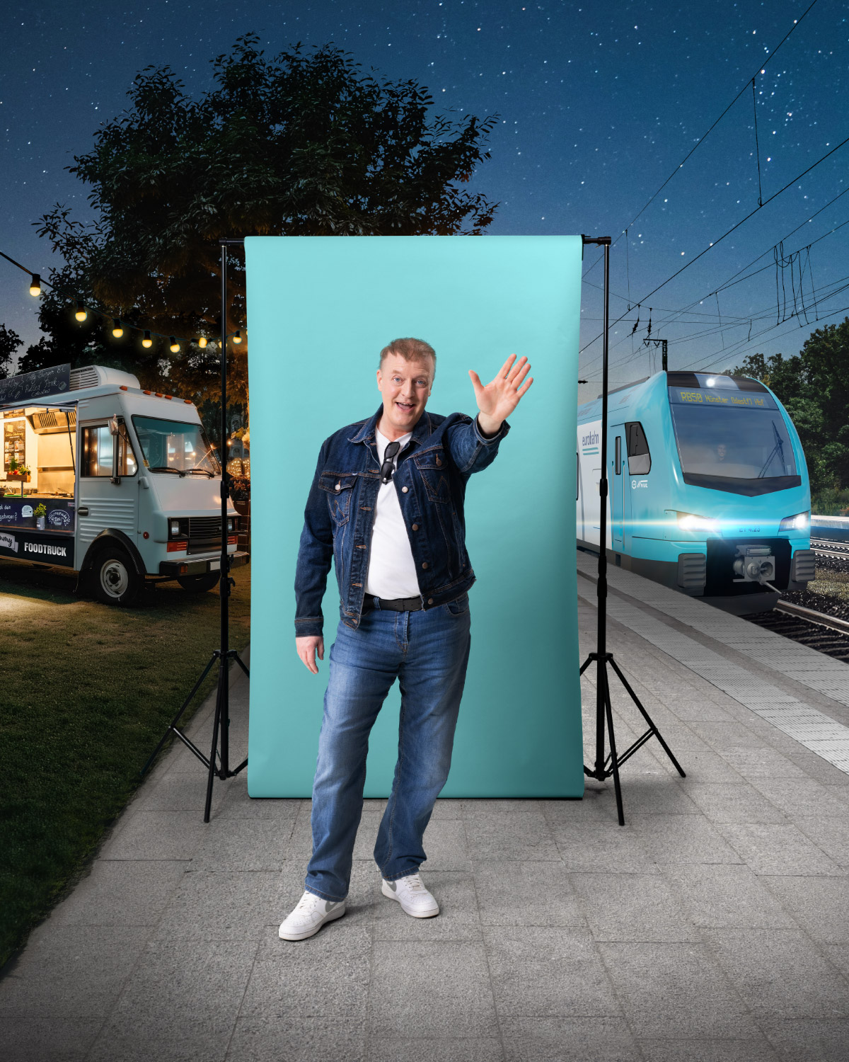 Ein mittelalter Mann in Jeansjacke und Jeans steht vor einer türkisen Leinwand und winkt mit einer Hand. Links ist ein beleuchteter Foodtruck zu sehen und rechts ein türkiser Zug der eurobahn. Die Szene findet an dem Bahnsteig Lünen bei Nacht statt unter einem sternenklaren Himmel.